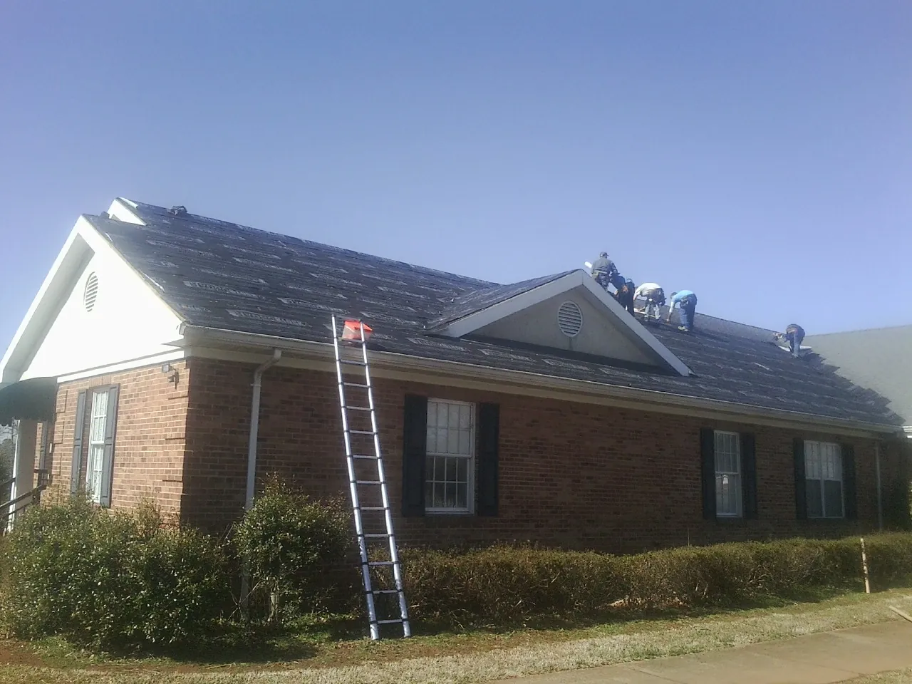 Crew tearing shingles off a brick ranch roof with a ladder set — Emergency Roof Repair work by our crew in Fort Drum