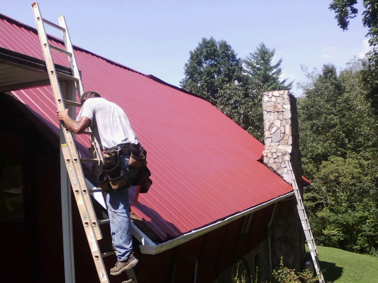 Roofer on a ladder beside a red metal roof and stone chimney — Soffit & Fascia Repair by Roofing Brothers in Fort Drum
